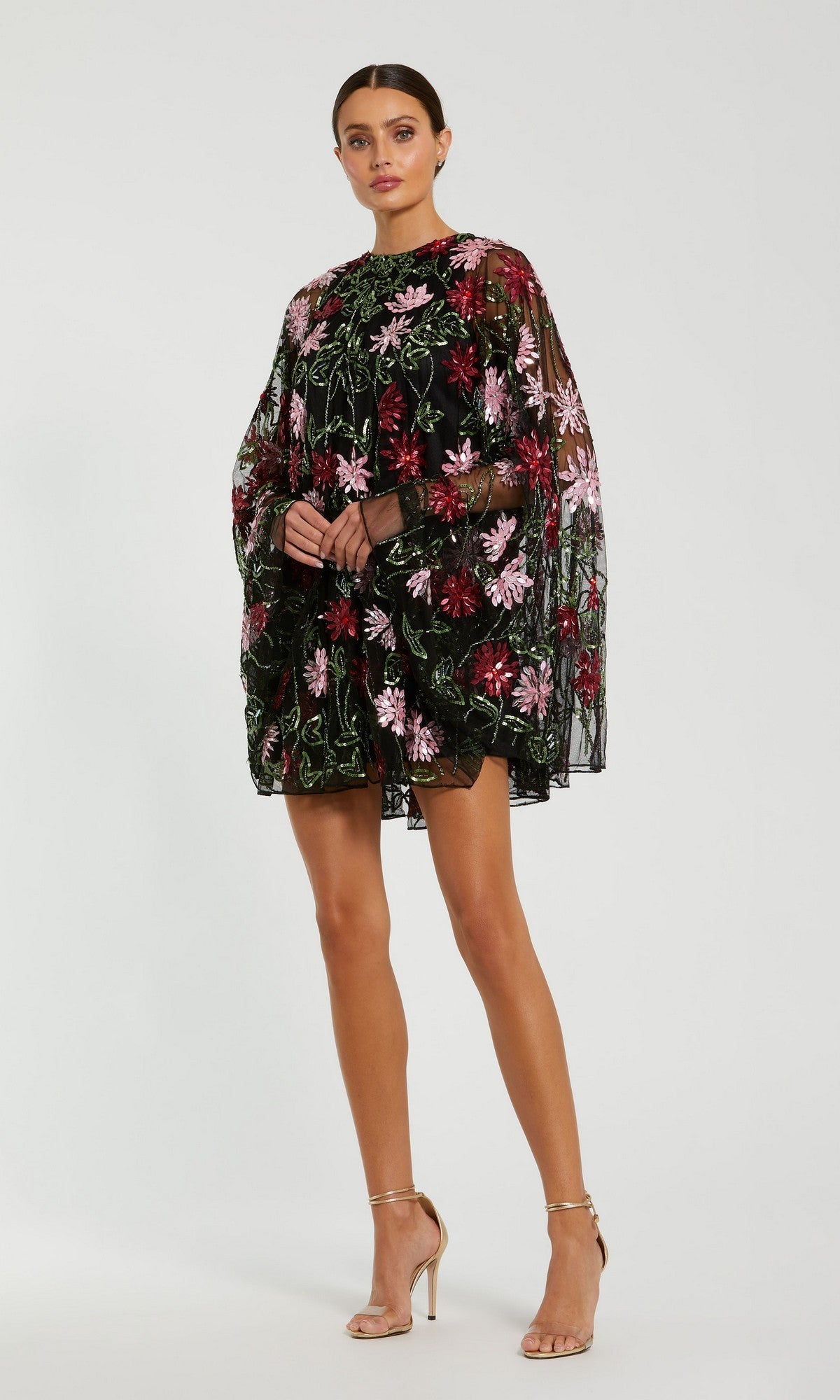 A woman stands against a plain light background in the Mac Duggal 6123 Sequin Short Wedding Guest Dress, a black sheer piece with pink and green floral embroidery, long sleeves, and beige high-heeled sandals that exude elegance.